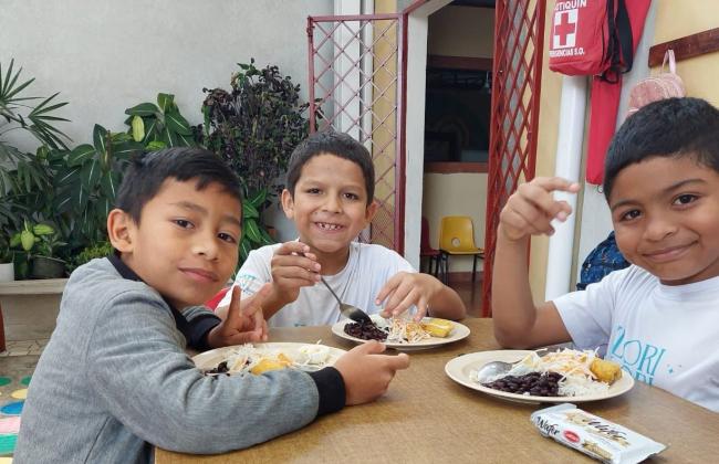Tres niños están sentados ante una mesa mientras sonríen y comen un plato de comida.