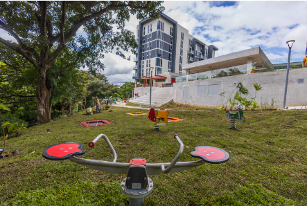 Fotografía de un parque de juegos en un espacio verde con árboles al lado izquierdo, al frente tiene un edificio residencial y una gradería.