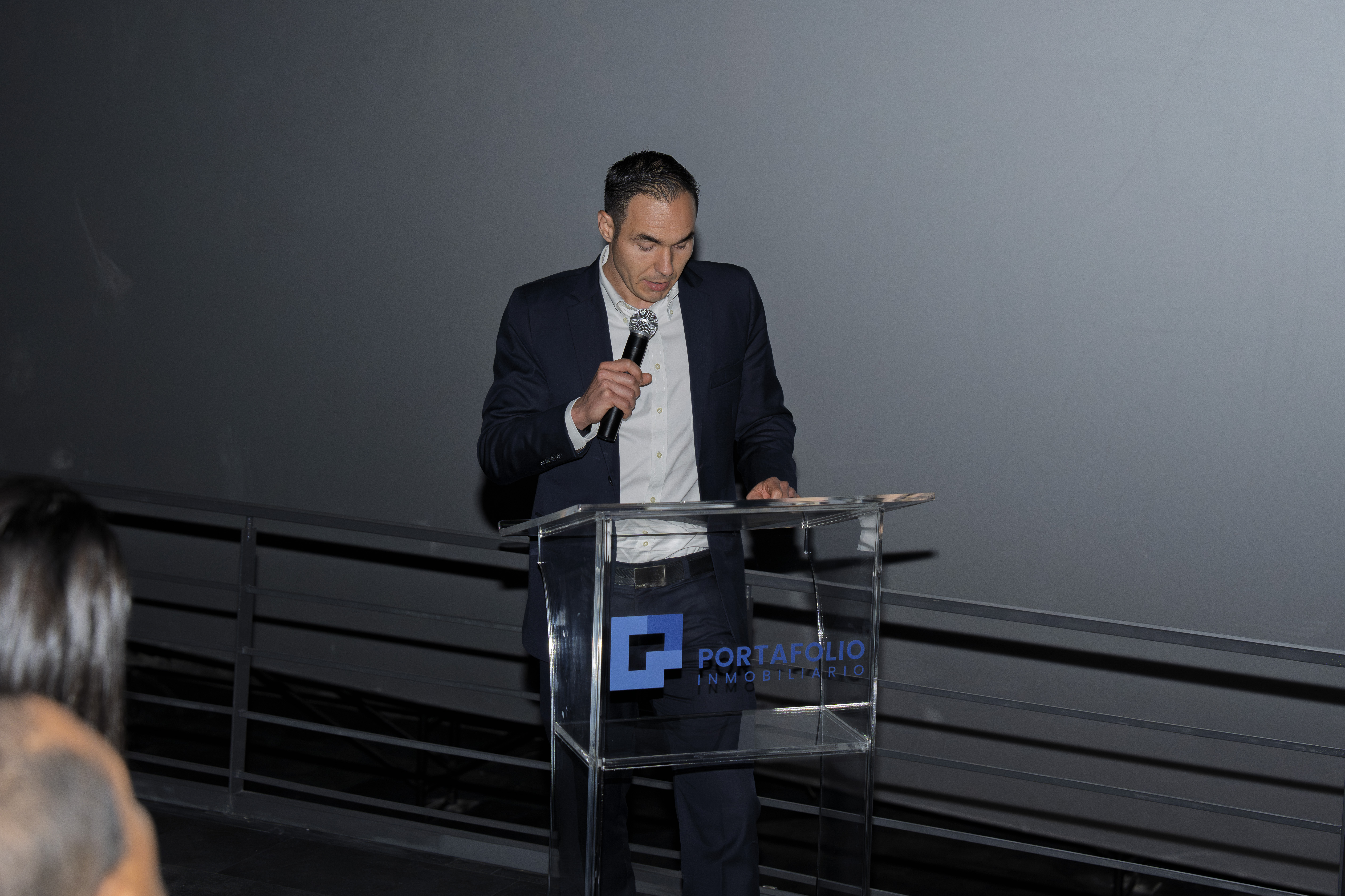 "Alfredo Volio director ejecutivo de la empresa viste de traje oscuro, camisa blanca y corbata se encuentra de pie detrás de un podio transparente con el logotipo de Portafolio Inmobiliario. Está sosteniendo un micrófono mientras parece dirigirse a una audiencia en un evento formal."