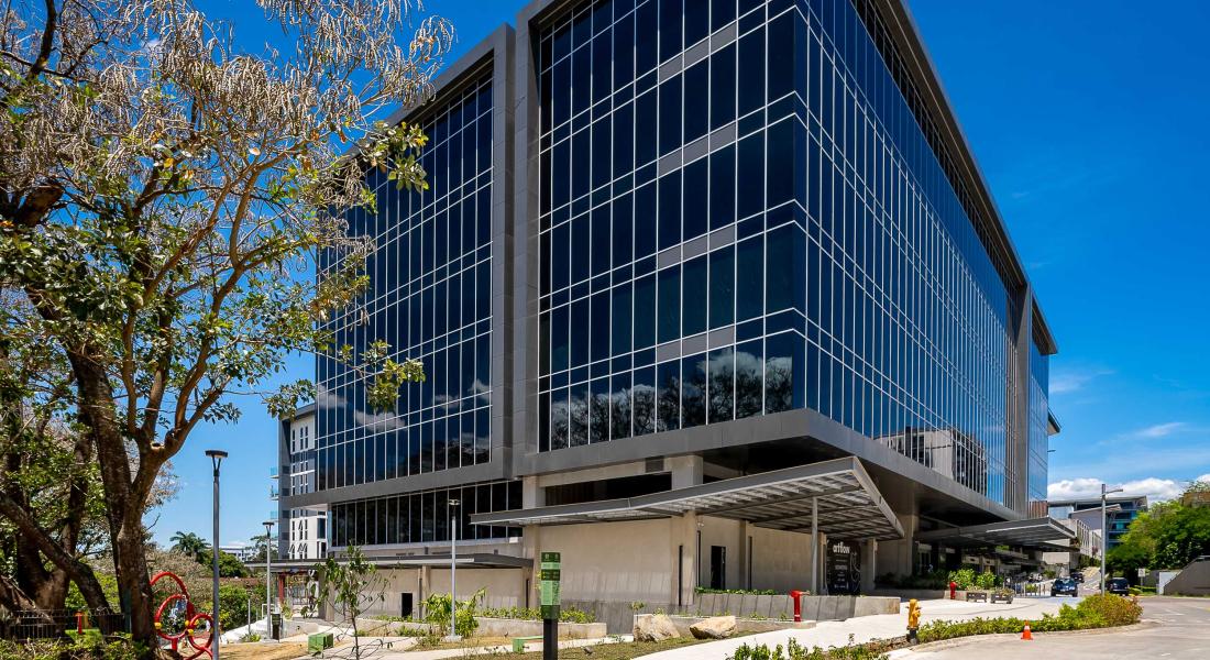 Edificio corporativo moderno de varios pisos con fachada de vidrio y estructura metálica, ubicado en un entorno urbano con aceras amplias, zonas verdes, árboles y mobiliario urbano como bancas y señalización. El cielo está despejado y azul, lo que resalta la arquitectura contemporánea del inmueble