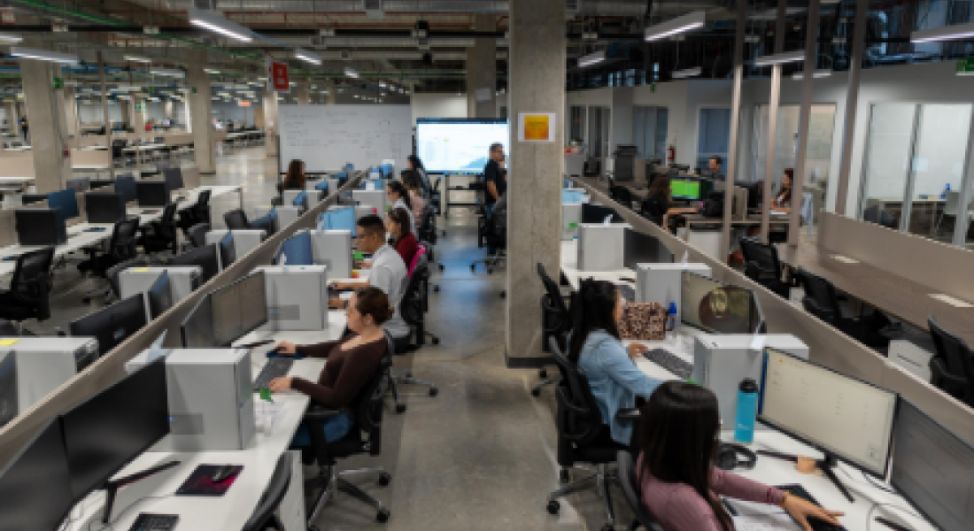 Amplio espacio de oficina con varias estaciones de trabajo en filas, donde numerosos empleados están sentados frente a sus computadoras. El diseño es de estilo abierto, con divisiones bajas entre los escritorios. Al fondo, una persona parece dar una presentación frente a una pantalla grande, y se observa un ambiente moderno, con columnas de concreto y tuberías expuestas en el techo. La oficina tiene un ambiente de actividad y colaboración.