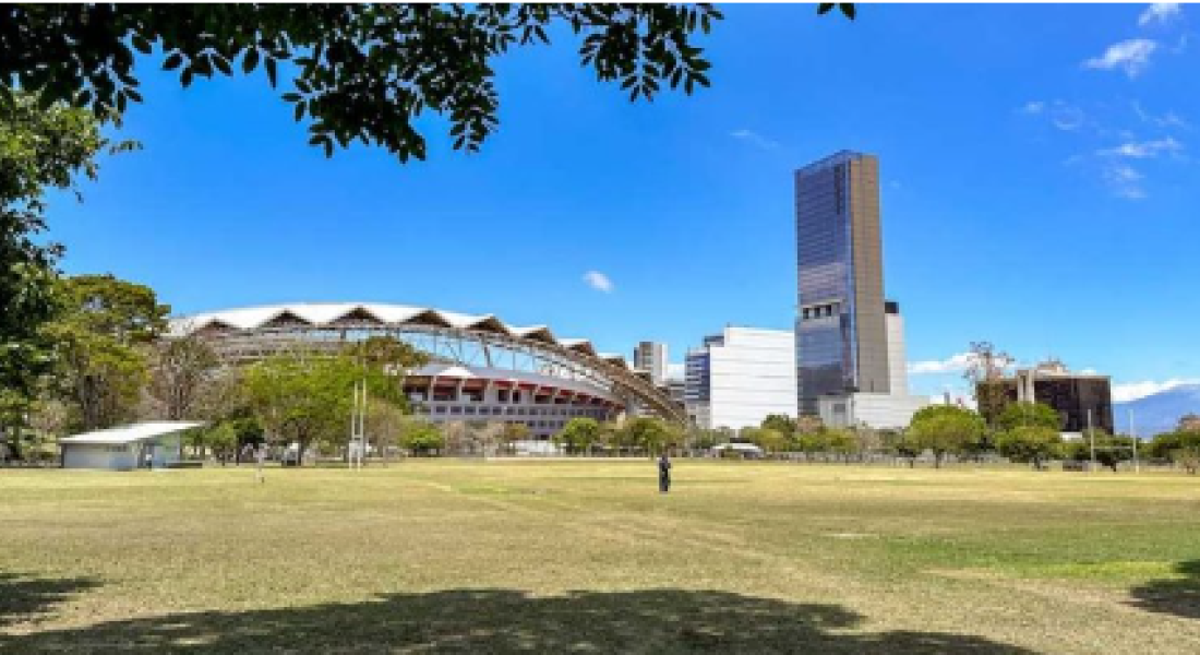 Amplio parque con áreas de césped y algunos árboles en primer plano, que ofrece una vista hacia el Estadio Nacional y un edificio alto y moderno en el horizonte. El cielo está despejado y azul, y se puede ver la estructura distintiva del estadio en contraste con el edificio de gran altura y otras edificaciones en la zona urbana. Un par de personas disfrutan del espacio abierto bajo la luz del sol.