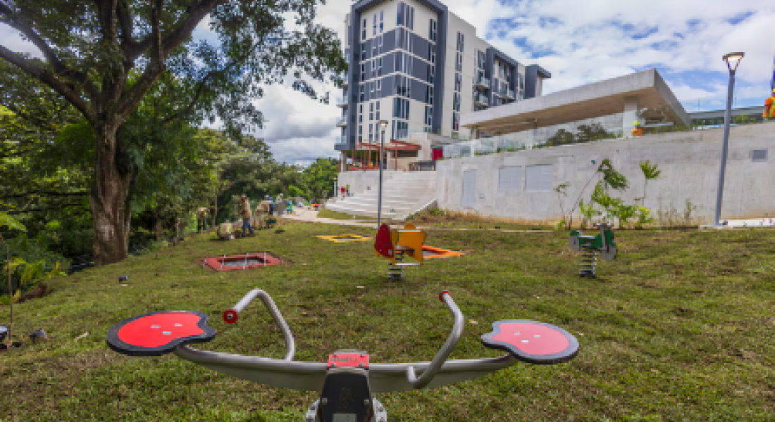 Fotografía de un parque de juegos en un espacio verde con árboles al lado izquierdo, al frente tiene un edificio residencial y una gradería.