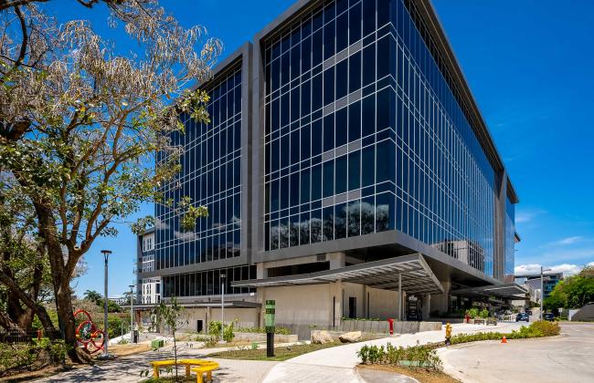 Edificio corporativo moderno de varios pisos con fachada de vidrio y estructura metálica, ubicado en un entorno urbano con aceras amplias, zonas verdes, árboles y mobiliario urbano como bancas y señalización. El cielo está despejado y azul, lo que resalta la arquitectura contemporánea del inmueble