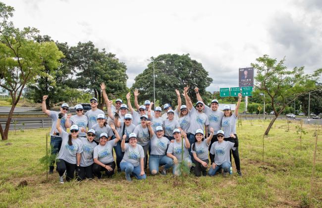 Un grupo de voluntarios vestidos con camisas grises posa en un terreno verde, en medio de pequeños árboles recién plantados.