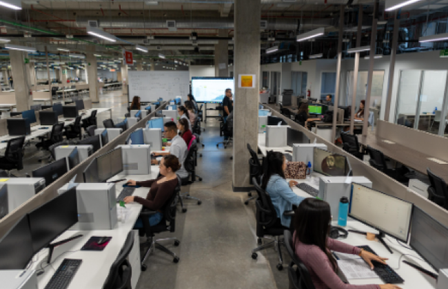 Amplio espacio de oficina con varias estaciones de trabajo en filas, donde numerosos empleados están sentados frente a sus computadoras. El diseño es de estilo abierto, con divisiones bajas entre los escritorios. Al fondo, una persona parece dar una presentación frente a una pantalla grande, y se observa un ambiente moderno, con columnas de concreto y tuberías expuestas en el techo. La oficina tiene un ambiente de actividad y colaboración.