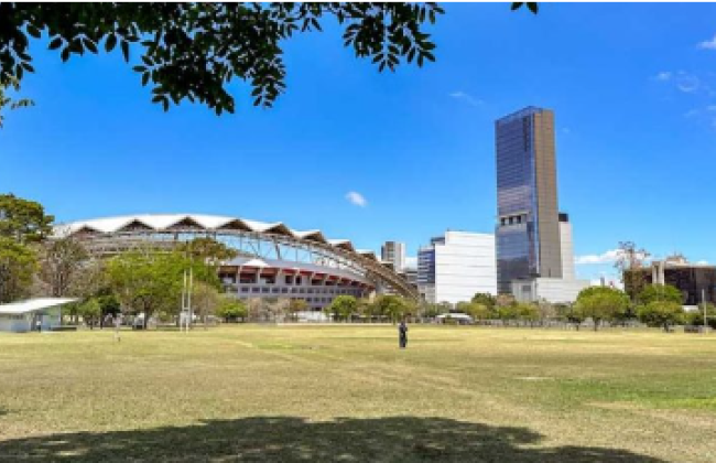 Amplio parque con áreas de césped y algunos árboles en primer plano, que ofrece una vista hacia el Estadio Nacional y un edificio alto y moderno en el horizonte. El cielo está despejado y azul, y se puede ver la estructura distintiva del estadio en contraste con el edificio de gran altura y otras edificaciones en la zona urbana. Un par de personas disfrutan del espacio abierto bajo la luz del sol.