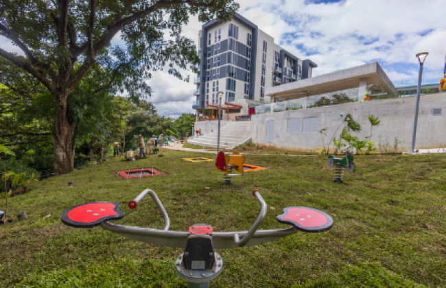 Fotografía de un parque de juegos en un espacio verde con árboles al lado izquierdo, al frente tiene un edificio residencial y una gradería.