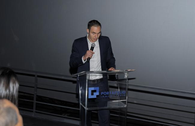 "Alfredo Volio director ejecutivo de la empresa viste de traje oscuro, camisa blanca y corbata se encuentra de pie detrás de un podio transparente con el logotipo de Portafolio Inmobiliario. Está sosteniendo un micrófono mientras parece dirigirse a una audiencia en un evento formal."