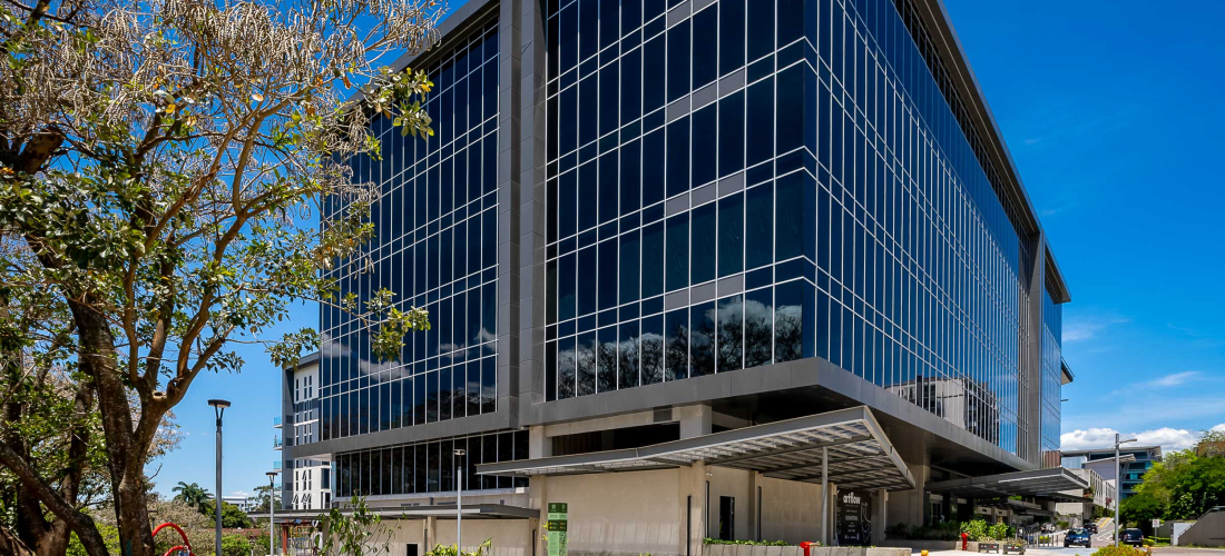 Edificio corporativo moderno de varios pisos con fachada de vidrio y estructura metálica, ubicado en un entorno urbano con aceras amplias, zonas verdes, árboles y mobiliario urbano como bancas y señalización. El cielo está despejado y azul, lo que resalta la arquitectura contemporánea del inmueble
