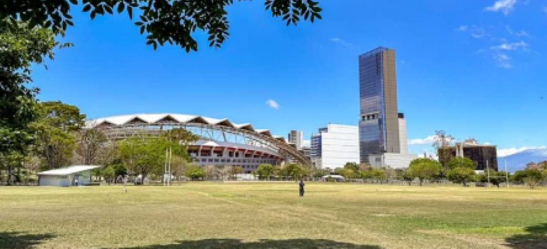 Amplio parque con áreas de césped y algunos árboles en primer plano, que ofrece una vista hacia el Estadio Nacional y un edificio alto y moderno en el horizonte. El cielo está despejado y azul, y se puede ver la estructura distintiva del estadio en contraste con el edificio de gran altura y otras edificaciones en la zona urbana. Un par de personas disfrutan del espacio abierto bajo la luz del sol.