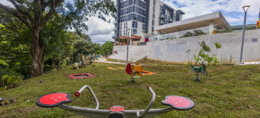 Fotografía de un parque de juegos en un espacio verde con árboles al lado izquierdo, al frente tiene un edificio residencial y una gradería.