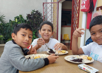 Tres niños están sentados ante una mesa mientras sonríen y comen un plato de comida.