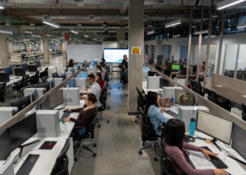 Amplio espacio de oficina con varias estaciones de trabajo en filas, donde numerosos empleados están sentados frente a sus computadoras. El diseño es de estilo abierto, con divisiones bajas entre los escritorios. Al fondo, una persona parece dar una presentación frente a una pantalla grande, y se observa un ambiente moderno, con columnas de concreto y tuberías expuestas en el techo. La oficina tiene un ambiente de actividad y colaboración.