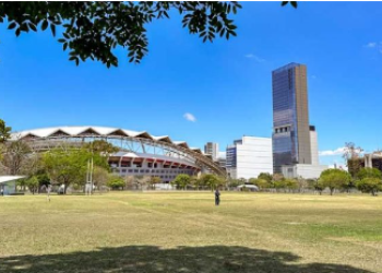 Amplio parque con áreas de césped y algunos árboles en primer plano, que ofrece una vista hacia el Estadio Nacional y un edificio alto y moderno en el horizonte. El cielo está despejado y azul, y se puede ver la estructura distintiva del estadio en contraste con el edificio de gran altura y otras edificaciones en la zona urbana. Un par de personas disfrutan del espacio abierto bajo la luz del sol.