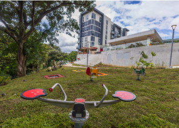 Fotografía de un parque de juegos en un espacio verde con árboles al lado izquierdo, al frente tiene un edificio residencial y una gradería.